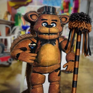 May include: A brown cardboard cutout of Freddy Fazbear, a character from the Five Nights at Freddy's video game series. He wears a black top hat and bow tie, and holds a microphone. A decorative staff with black and orange accents is to the right.