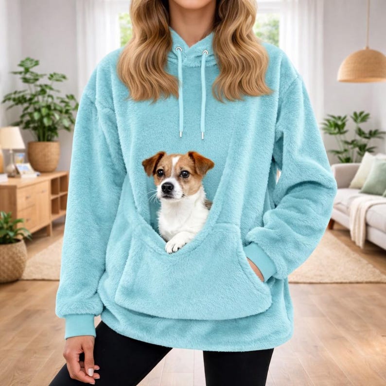 May include: A light blue, fluffy hoodie with a kangaroo pocket, holding a small dog. The dog has brown and white fur. The hoodie has a drawstring hood and ribbed cuffs. The person is wearing black leggings.