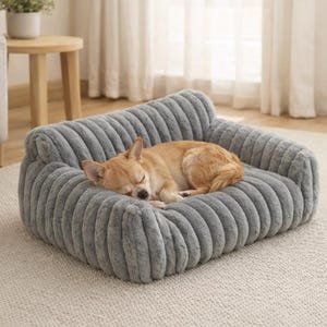 May include: A small, light brown dog asleep on a grey, plush dog bed. The bed is shaped like a miniature sofa with a ribbed texture. The dog is curled up, appearing comfortable and relaxed.