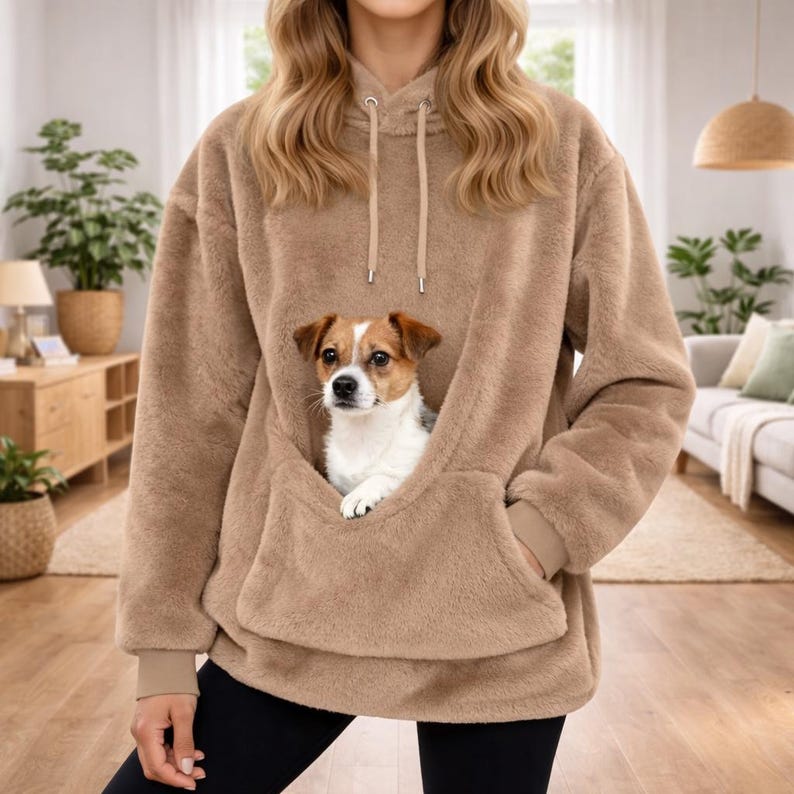 May include: A tan, oversized hoodie with a large front pocket, featuring a small dog nestled inside. The hoodie has a drawstring hood and ribbed cuffs. The dog is a Jack Russell Terrier mix with brown, white, and tan markings. The hoodie is made of a soft, plush material.