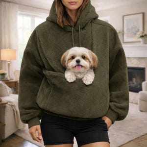 May include: An olive green fuzzy fleece hoodie featuring a large front pouch pocket. A small, long-haired white and tan dog with floppy ears and a pink tongue is positioned inside the pouch, with its front paws resting on the top edge of the pocket.