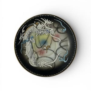 May include: A small, round, black dish featuring a white dragon design. The dragon is intricately drawn with a long body and detailed scales. The dish has a gold rim and a colourful, abstract background.