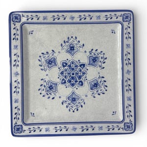 May include: Square ceramic plate with a white background and a blue floral design. The plate has a blue border with floral and leaf patterns. The center features a large blue floral motif, creating a decorative and elegant look.
