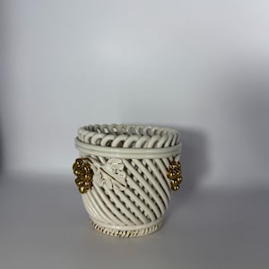 May include: Cream-coloured ceramic basket-style vase with a woven design. The vase has gold-coloured grape cluster and leaf accents. The top rim has an openwork design. The base has a gold-coloured trim.