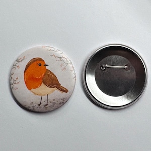 Könnte beinhalten: Ein runder Button mit einer Aquarellillustration eines Rotkehlchens. Der Vogel hat eine orangefarbene Brust, braune Flügel und ist vor einem weichen, weißen Hintergrund mit zarten floralen Akzenten platziert. Die Rückseite des Buttons ist silberfarben.