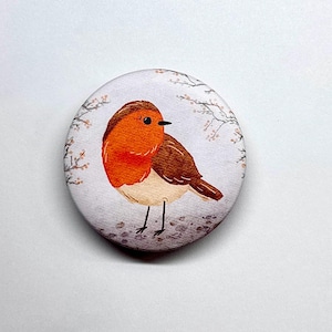 Könnte beinhalten: Ein runder Button mit einer Aquarellillustration eines Rotkehlchens. Der Vogel hat eine leuchtend orangefarbene Brust, braune Flügel und ist vor einem weißen Hintergrund mit zarten floralen Akzenten platziert. Ein bezauberndes Accessoire.