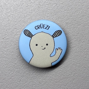 Könnte beinhalten: Ein runder, hellblauer Button mit einer Cartoon-Illustration eines freundlichen, hellgrauen Wesens, das winkt. Der Button hat das Wort "GRÜEZI" in schwarzem Text.