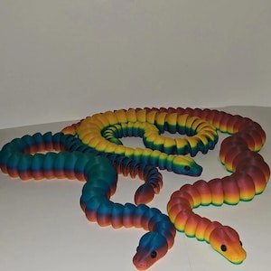 May include: Several colorful, articulated snake toys in shades of red, orange, yellow, green, blue, and purple. The snakes are posed on a white surface, with their segmented bodies curved and their heads raised. Each snake has a small, rounded head.
