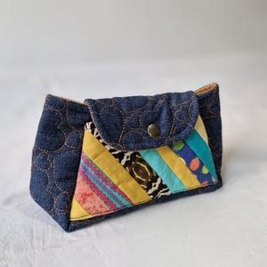 May include: A patchwork wallet made from denim. The flap is secured with a metal popper. The patchwork incorporates fabrics in yellow, pink, blue, and animal print patterns. The denim has a quilted heart design.