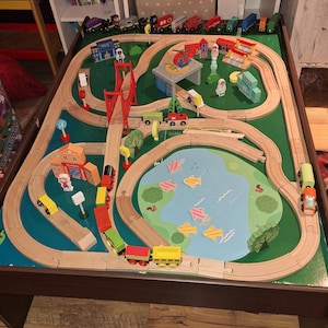 May include: Wooden train set with a green play surface, featuring a bridge, buildings, a lake with fish, and a helicopter landing pad. The set includes various colorful train cars and accessories for imaginative play.
