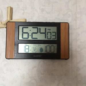 May include: Digital atomic clock with a black frame and wood-grain accents. The display shows the time as 6:24 PM, the date, and the outdoor temperature of 100 degrees Fahrenheit. The clock also displays the moon phase.