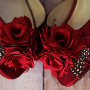 May include: A pair of red satin shoes with a large red rose embellishment on each toe. The roses are made of fabric and have a ruffled texture. The shoes also have a small feather accent on each side.