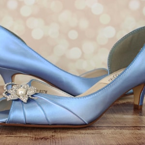 May include: A pair of light blue satin peep-toe heels with a rhinestone flower embellishment. The heels are approximately 2.5 inches tall.