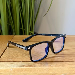 May include: Black-framed eyeglasses with a rectangular shape and blue accents. The temples feature decorative silver embellishments and a cross design. The lenses have a light tint. The glasses are displayed on a wooden surface.