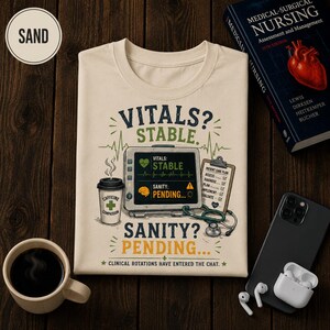 Könnte beinhalten: Ein sandfarbenes T-Shirt mit einer Grafik eines medizinischen Monitors, der Vitals Stable und Sanity Pending anzeigt. Das Design umfasst einen Kaffeebecher mit der Aufschrift Caffeine Compassion, ein Klemmbrett, ein Stethoskop und den Text Clinical Rotations Have Entered The Chat.