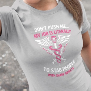 May include: A pale grey t-shirt featuring the text "DON'T PUSH ME... MY JOB IS LITERALLY TO STAB PEOPLE WITH SHARP OBJECTS." The design includes a medical symbol, wings, syringes, and a snake.