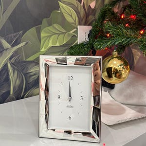 Może przedstawiać: Srebrny zegar z prostokątną ramą. Tarcza zegara jest biała z czarnymi cyframi i wskazówkami, wskazującymi godzinę 12:00. Na tarczy zegara wydrukowano słowo "atelier". Zegar stoi na białej powierzchni.