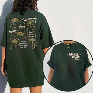 May include: Dark green t-shirt with a humorous design. The back features cartoon UFOs and text including "FIST MY BUMP!" and "SARCASM...". The front displays "AMAZE!" in white. A fun, casual apparel item.
