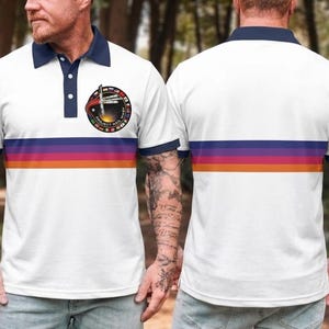 May include: White polo shirt with a navy blue collar and trim, displaying a horizontal stripe in purple, orange, and red. The shirt has a circular emblem with text and a graphic. The back of the shirt also has the horizontal stripe.