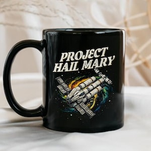 Puede incluir: Taza de cerámica negra con el texto blanco "PROJECT HAIL MARY" y una ilustración de una nave espacial sobre un fondo de nebulosa colorida. La taza tiene un asa curva.