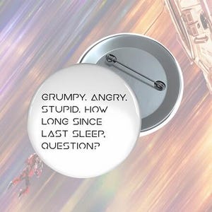 May include: A white button with the text "GRUMPY. ANGRY. STUPID. HOW LONG SINCE LAST SLEEP, QUESTION?" in a black, futuristic font. The button is round with a metal pin back. The background is a blurred mix of colors.