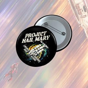 May include: A black button with the text "PROJECT HAIL MARY" in white, featuring a spaceship design against a colorful galaxy backdrop. A second, silver button is partially visible. The design is science fiction themed.