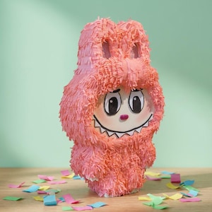 Può includere: Una piñata rosa a forma di mostro dei cartoni animati con un volto sorridente, grandi occhi e un sorriso con denti aguzzi. La piñata è ricoperta di carta frangiata e poggia su una superficie di legno con coriandoli colorati sparsi.