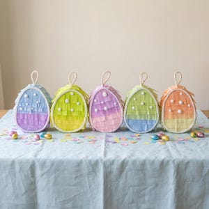Può includere: Cinque piñatas colorate a forma di uovo di Pasqua con bordi bianchi e manici in corda. Ogni piñata è decorata con strati di carta frangiata nelle tonalità del blu, verde, rosa e arancione. Piccoli punti bianchi sono sparsi sul davanti e coriandoli li circondano.