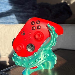 May include: A red Xbox controller rests on a teal and blue tentacle-shaped stand. The controller has black buttons and a black directional pad. The stand is 3D-printed and has a textured surface, with the tentacles curling upwards.
