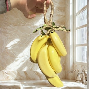 May include: A bunch of yellow banana-shaped decorative items, held by a rope loop. The bananas are clustered together and tied with twine and green leaves. The background shows a marble-like surface.