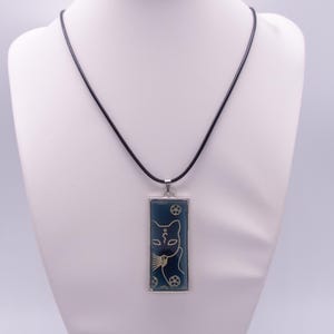 Peut inclure: Un collier pendentif rectangulaire avec un fond bleu foncé orné d'une illustration au trait doré représentant un visage de chat et de petites fleurs. Le pendentif est entouré d'un cadre en métal argenté et suspendu à un cordon noir.