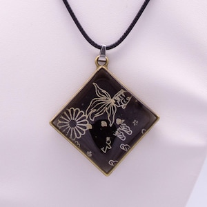 Puede incluir: Un colgante en forma de diamante con fondo negro y dibujos lineales en color dorado de un pez dorado y una flor. El colgante está engastado en un marco de metal color bronce y cuelga de un sencillo cordón negro.