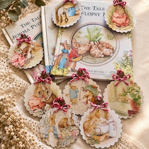 May include: Eight round wooden ornaments featuring Beatrix Potter illustrations of rabbits, each topped with a red and white twine bow. The ornaments are displayed over two books titled The Tale of Jemima Puddle-Duck and The Tale of the Flopsy Bunnies.