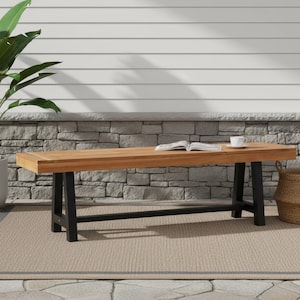 May include: A wooden outdoor bench with black legs, a light brown tabletop, and a woven basket. A book and a white cup sit on the bench. A large potted plant is to the left, and the bench sits on a beige rug.