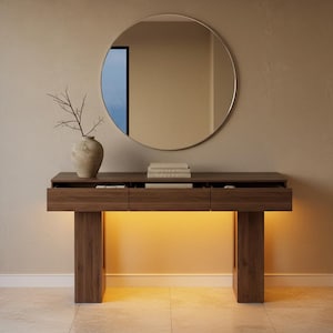 May include: A modern wooden console table with three drawers and a warm under-glow. A round mirror hangs above the table, reflecting the room. A vase with branches and a stack of books are on the table.