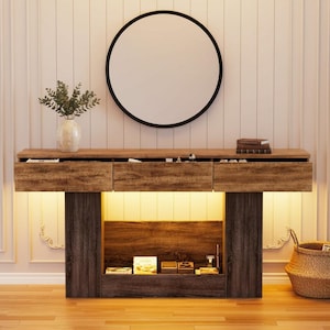 May include: A wooden console table with three drawers, a round black-framed mirror, and a vase of greenery. The table has a dark wood base and a light wood top. A woven basket and books are also visible.