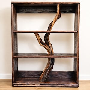 May include: A dark brown wooden bookshelf featuring three horizontal shelves and a central vertical support made from a natural, twisted tree branch that extends through the middle of the shelves, creating a unique structural design element.