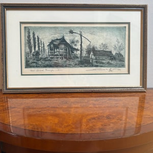 May include: A framed black and white etching depicting a rural landscape with a wooden structure, a tall well sweep, and trees. The artwork features a cream-coloured mat, a dark textured frame, and handwritten text at the bottom left and right.