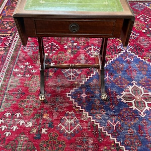 May include: A dark wood drop-leaf side table featuring a rectangular top with a green leather inlay, a single central drawer with a circular metal pull, and four legs ending in brass claw-and-ball feet with small casters for mobility.
