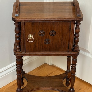 May include: A dark brown wooden side table featuring a rectangular top with curved side handles, a single cabinet door with a gold-toned metal ring pull, three circular carved accents, and four turned legs connected by a lower shelf.