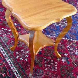 May include: A light brown wooden side table featuring a square top with curved edges and corners. The table is supported by four slender, curved legs that connect to a smaller, matching square lower shelf positioned between the legs.
