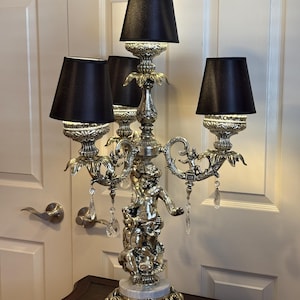 May include: A silver-toned candelabra lamp featuring a central cherub figure, three arms with black fabric shades, and hanging clear crystal teardrop accents. The lamp base is circular and rests on ornate feet, with a white marble-like accent piece.