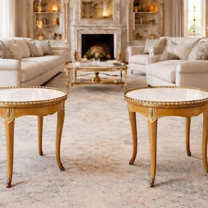 May include: A pair of round side tables featuring white marble tops and light wood frames with gold-toned metal trim along the edges. Each table stands on four curved legs with decorative gold-toned accents at the top of each leg.