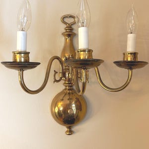 May include: A polished brass wall sconce featuring three curved arms extending from a central vertical mount. Each arm holds a circular drip pan and a white candle-style socket with a clear, flame-shaped light bulb. The fixture has a rounded base.