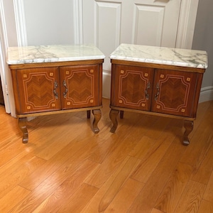 May include: A pair of wooden bedside cabinets featuring white marble tops with grey veining. Each cabinet has two doors with chevron wood grain patterns, light wood inlay borders, and ornate metal handles, supported by four curved legs.