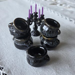 Peut inclure: Un ensemble de cinq chaudrons miniatures en céramique noire avec une finition brillante et un léger moucheté blanc, disposés en groupe. Derrière les chaudrons se trouve un petit candélabre noir portant cinq fines bougies violettes pailletées.