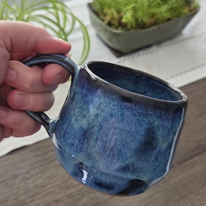 Peut inclure: Une tasse en céramique avec un vernis brillant et tacheté dans des tons bleu marine profond, bleu roi et bleu ciel plus clair. La tasse présente un corps arrondi légèrement effilé avec une anse épaisse et incurvée sur le côté, finition lisse.
