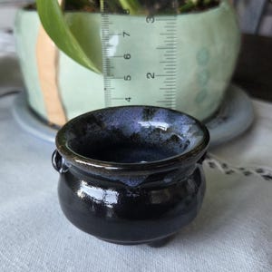 May include: A small, round, black ceramic cauldron with a glossy finish and a purple-tinted rim. A clear plastic ruler is positioned behind the pot, showing it stands approximately 3.8 cm tall from the base to the top rim.