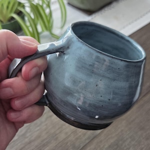 May include: A round ceramic mug with a glossy light blue glaze featuring subtle dark grey speckles and a darker, solid grey base. The mug has a thick, curved handle and a wide opening, showcasing a hand-thrown appearance with visible texture.
