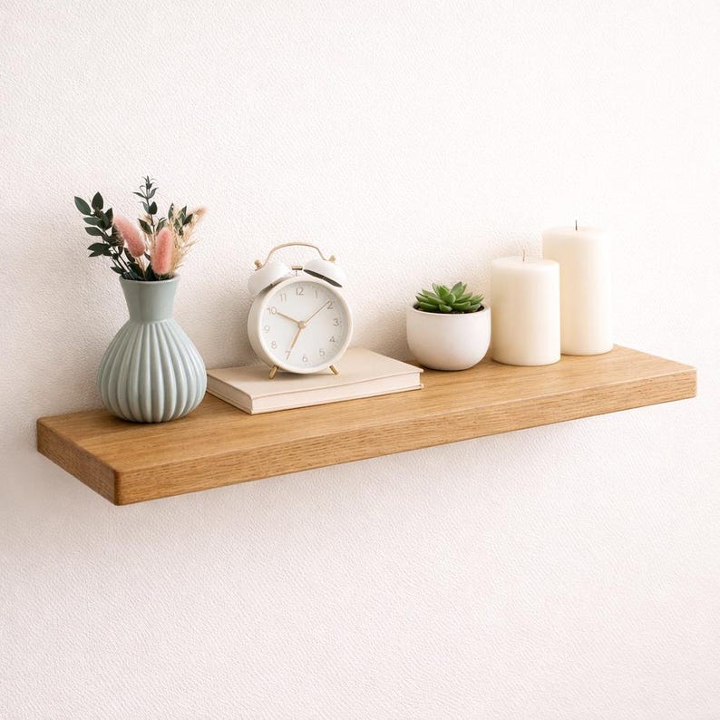 Puede incluir: Una estanter&iacute;a flotante de madera exhibe un peque&ntilde;o jarr&oacute;n con flores secas, un despertador blanco, una suculenta en una maceta blanca y dos velas de pilar blancas. La estanter&iacute;a es de madera clara.